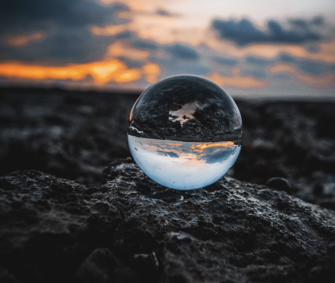Circular water trop resting on a rock with a sunset behind it, but in the drop, there is a reversed image of sky and the ground